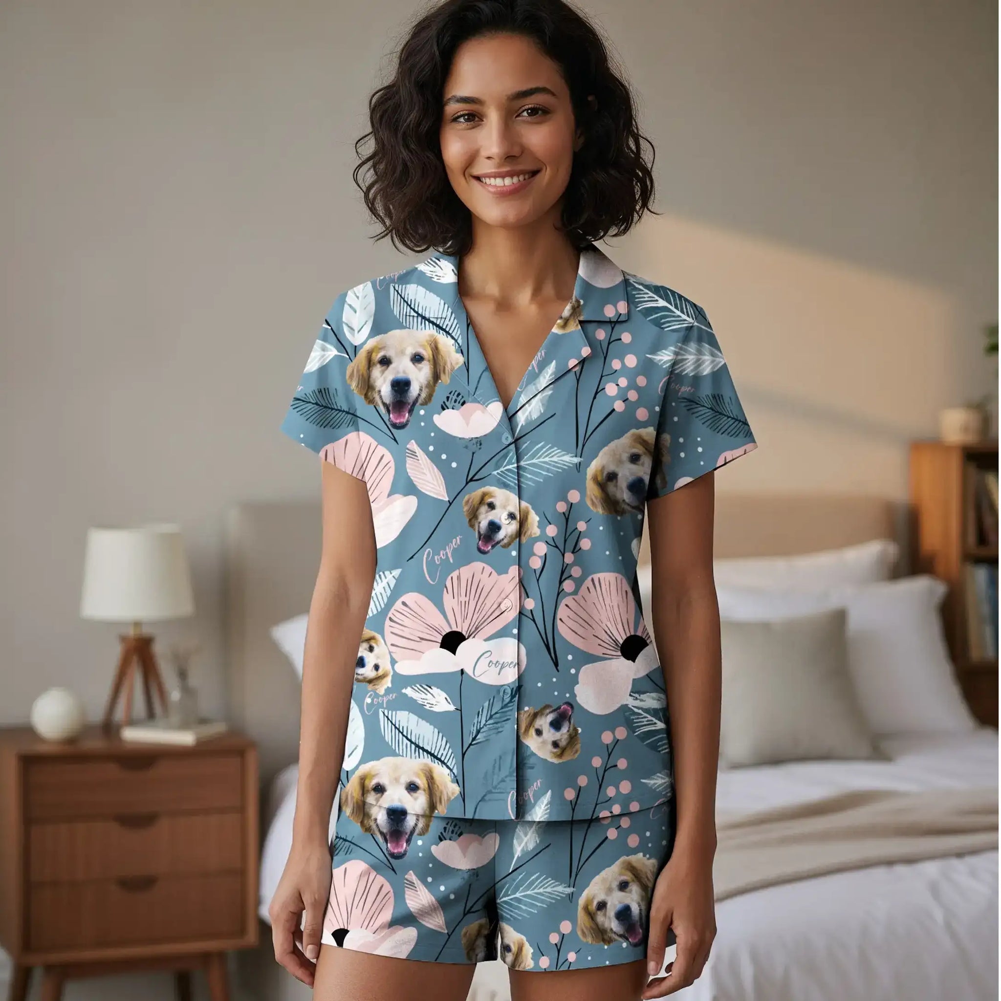 Photograph of a smiling, curly-haired woman in a blue, dog-patterned pajama set standing in a beige bedroom with a wooden nightstand.