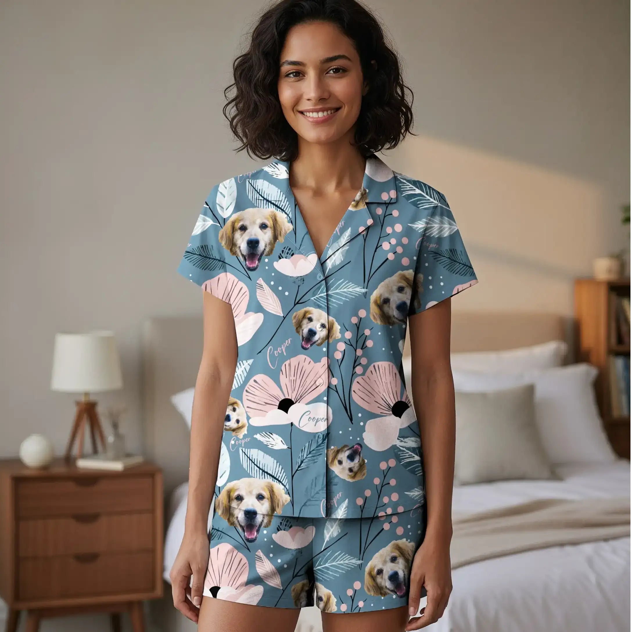 Photograph of a smiling, curly-haired woman in a blue, dog-patterned pajama set standing in a beige bedroom with a wooden nightstand.