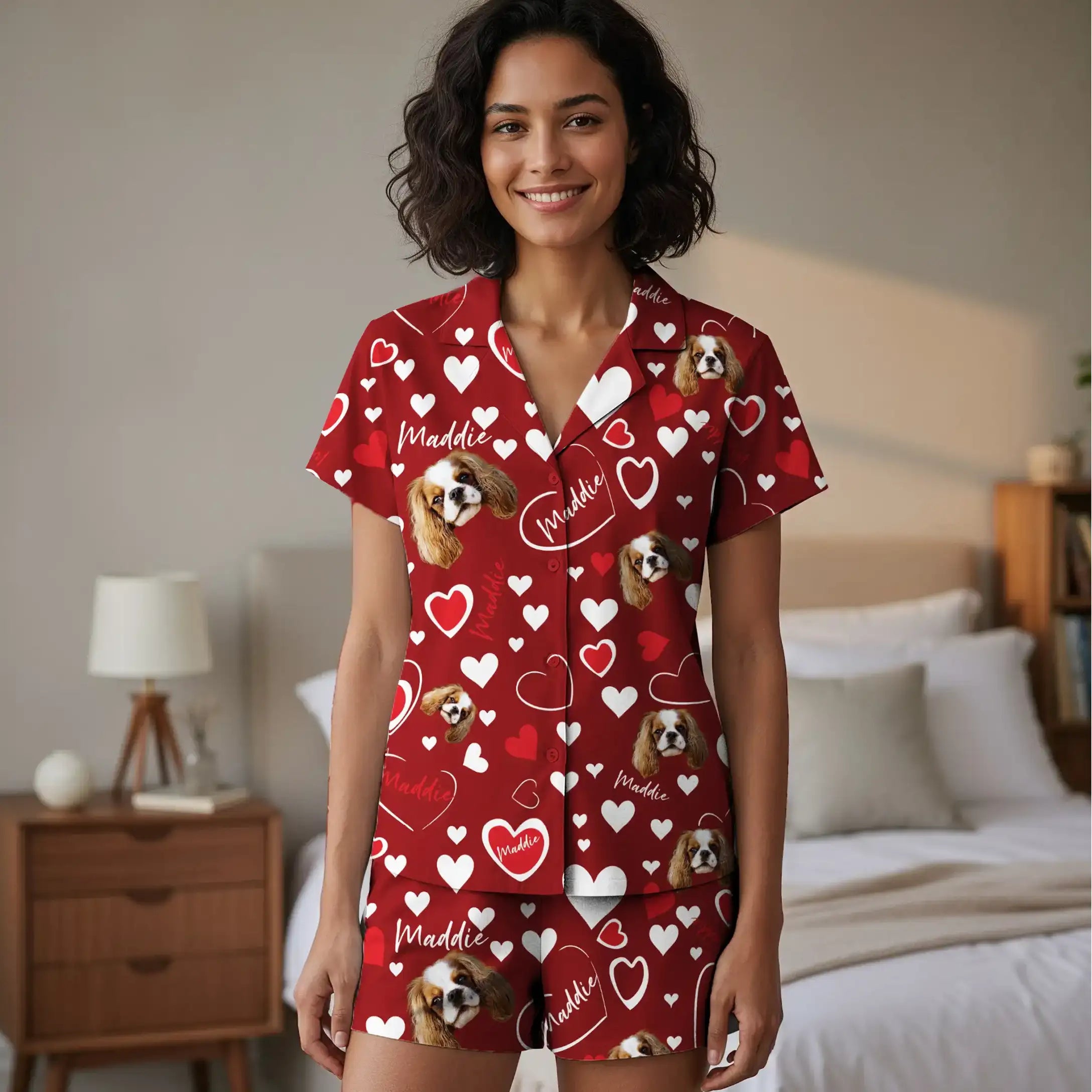 Photograph of a smiling, curly-haired woman in red pajamas with heart and dog patterns, standing in a beige bedroom.