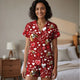Photograph of a smiling, curly-haired woman in red pajamas with heart and dog patterns, standing in a beige bedroom.