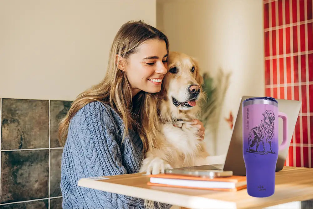 Photograph of a smiling young woman with long blonde hair, wearing a blue cable-knit sweater, hugging a golden retriever. They stand beside a wooden table with a purple travel mug, a laptop, and books. Background features tiled walls and red patterned wallpaper.